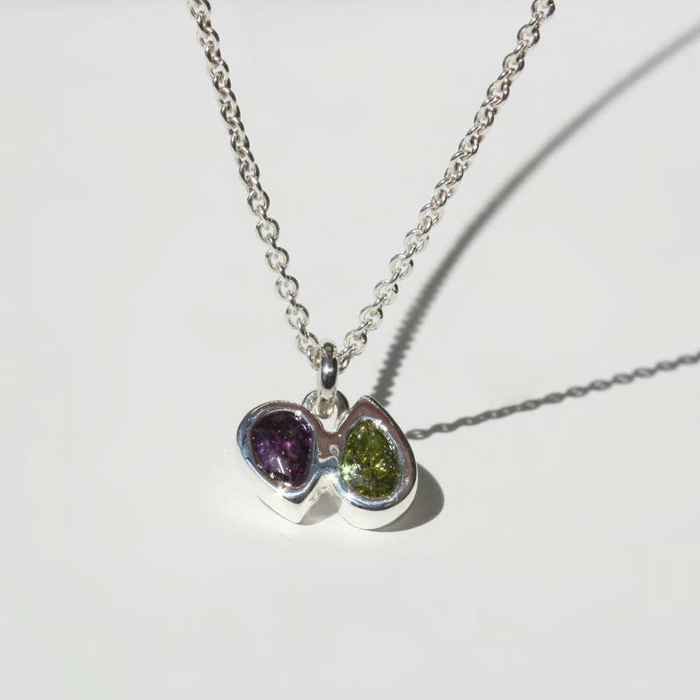 A silver chain necklace with a double-pear pendant featuring one purple gemstone and one green gemstone pear, set against a clean white background.