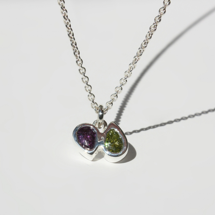 A silver chain necklace with a double-pear pendant featuring one purple gemstone and one green gemstone pear, set against a clean white background.