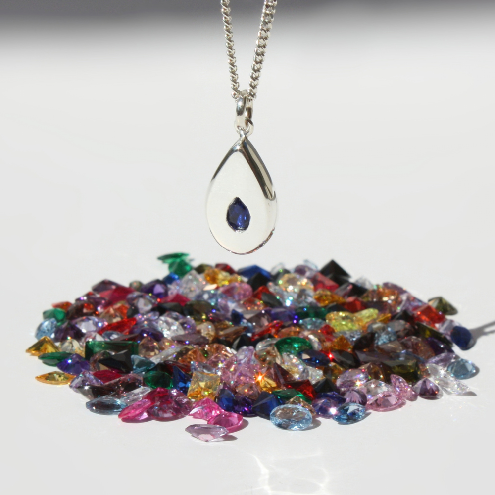 A silver teardrop pendant necklace with a small blue teardrop gemstone, suspended above a pile of many colorful loose gemstones on a white surface.
