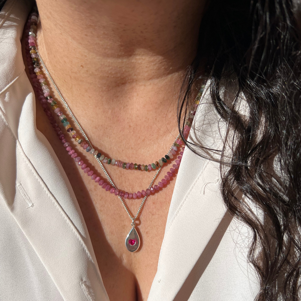 Close-up of a person wearing layered necklaces, including a silver chain with a teardrop pendant featuring a pink gemstone and a pink beaded necklace layered over a multicolor beaded strand.