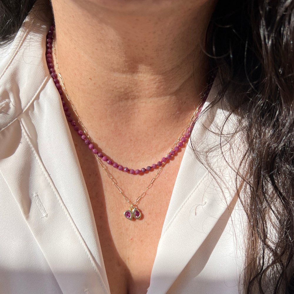 A close-up of a person wearing layered necklaces: a short beaded Ruby necklace and a 14k gold paperclip chain with a Fuchsia double pear necklace 