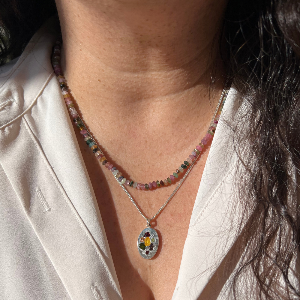 A close-up of a person wearing a layered necklace set. The top necklace is made of small multicolored faceted beads, and the bottom necklace is a silver chain with an oval pendant featuring a colorful gemstone design.