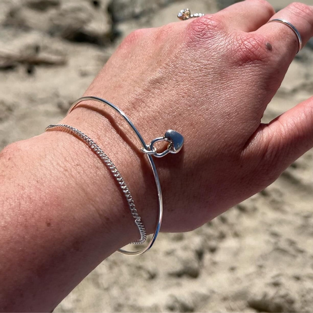 Model wearing a sterling silver heart charm bangle bracelet layered in a silver bangle stack.